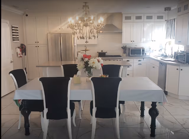White kitchen with a dining table and six black-and-white chairs, a chandelier overhead, and stainless-steel appliances.