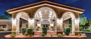 Front exterior view of Springdale Village Assisted Living Facility & Independent Living Apartments building at night, featuring a large covered entrance with arches, illuminated by warm lights, surrounded by neatly trimmed bushes and landscaping.