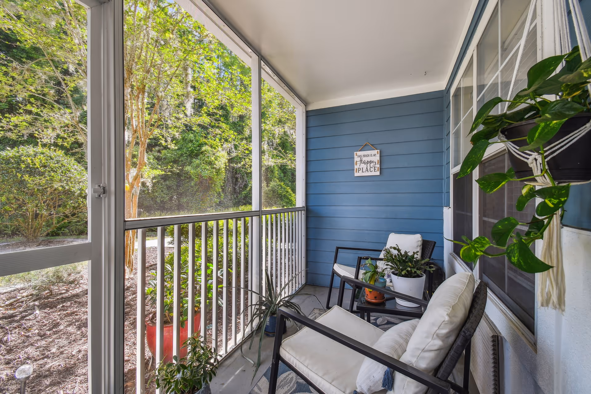 A cozy screened-in porch with two cushioned chairs, a small table with potted plants, and additional hanging and floor plants. The porch has blue siding on one wall and overlooks a green, wooded outdoor area.