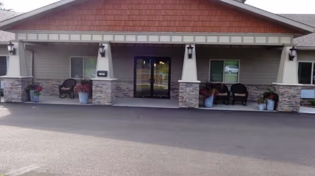 Front entrance of a single-story assisted living facility with a covered porch, double glass doors, stone-clad columns, chairs, and potted plants.