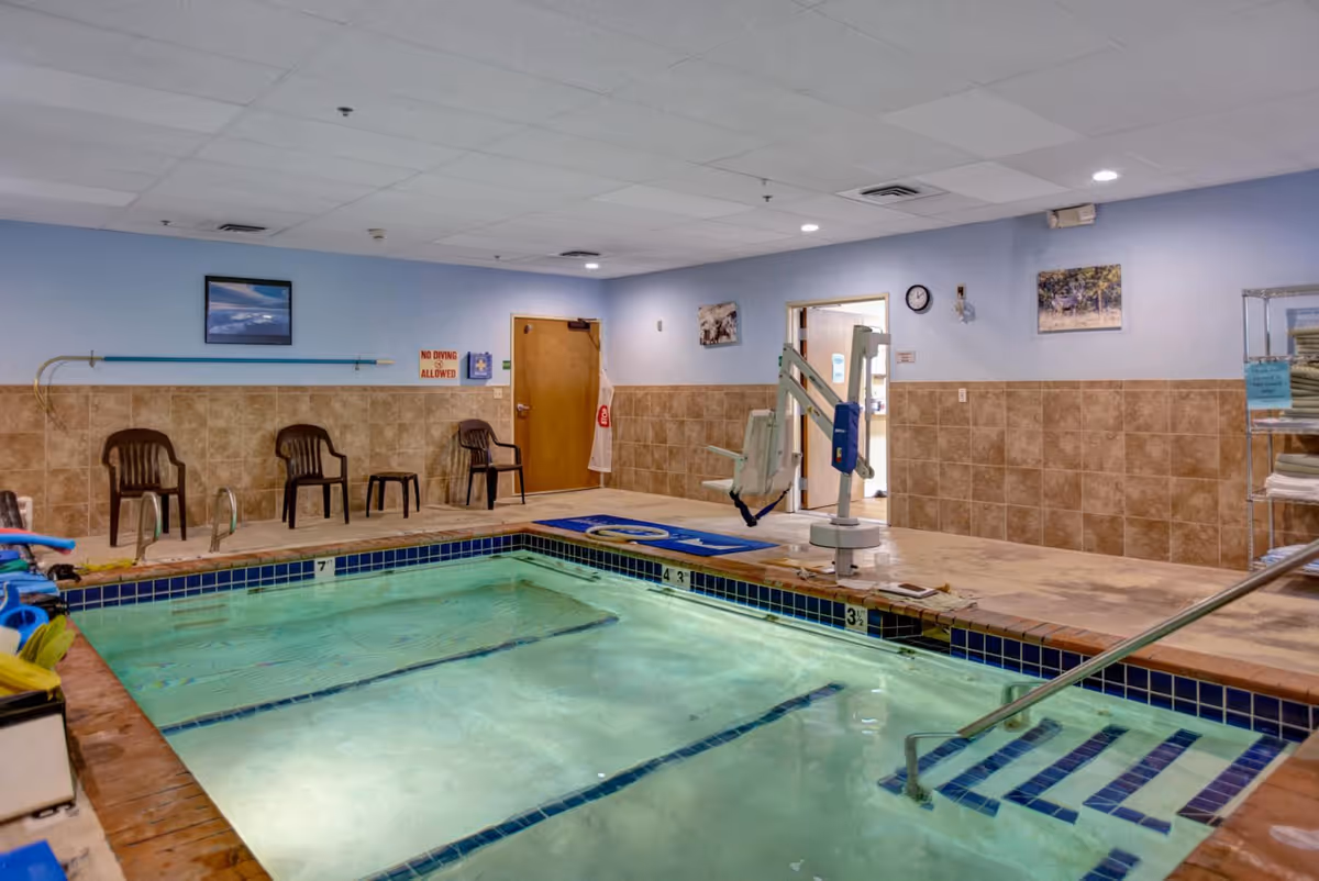 Indoor swimming pool area with clear water, tiled walls, and a ceiling with recessed lighting. There are several plastic chairs along the wall, a pool lift for accessibility, a metal handrail leading into the pool, and a rack with folded towels. The walls are decorated with framed pictures and a clock.