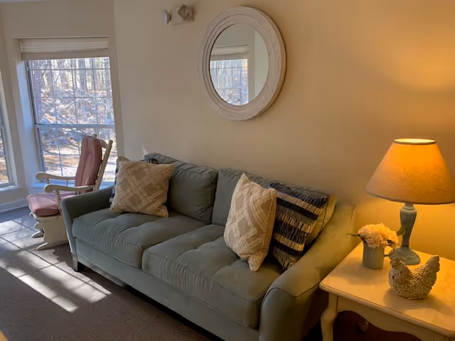 A cozy living room area with a light green sofa adorned with three decorative pillows. To the right of the sofa is a small white side table holding a lamp with a beige shade, a small flower arrangement, and a decorative chicken figurine. A round mirror hangs on the wall above the sofa. To the left, there is a large window with blinds partially open, and a cushioned rocking chair is positioned near the window.
