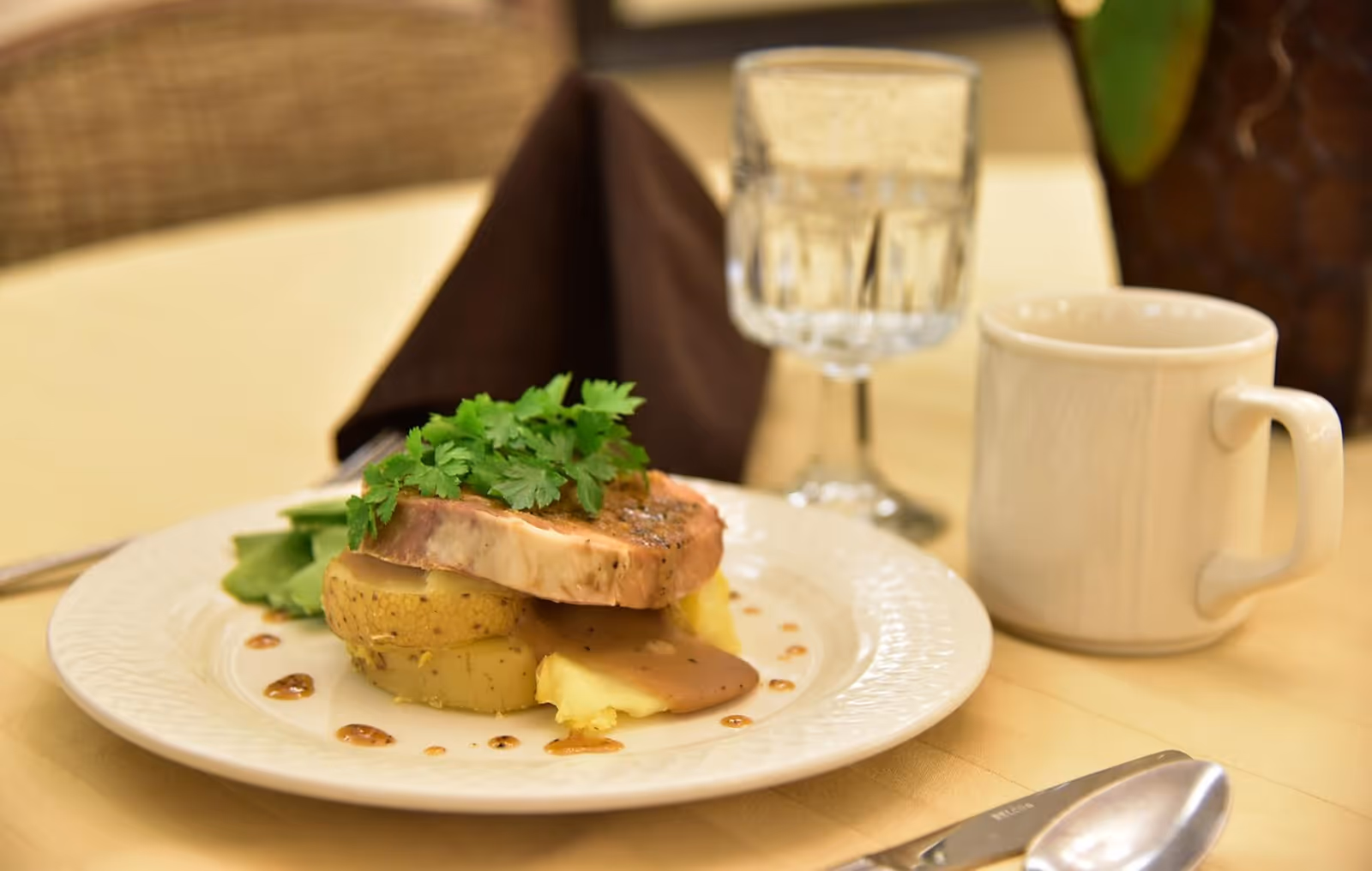 Plated entrée of meat and potatoes with sauce and parsley on a table next to a coffee mug and a water glass.