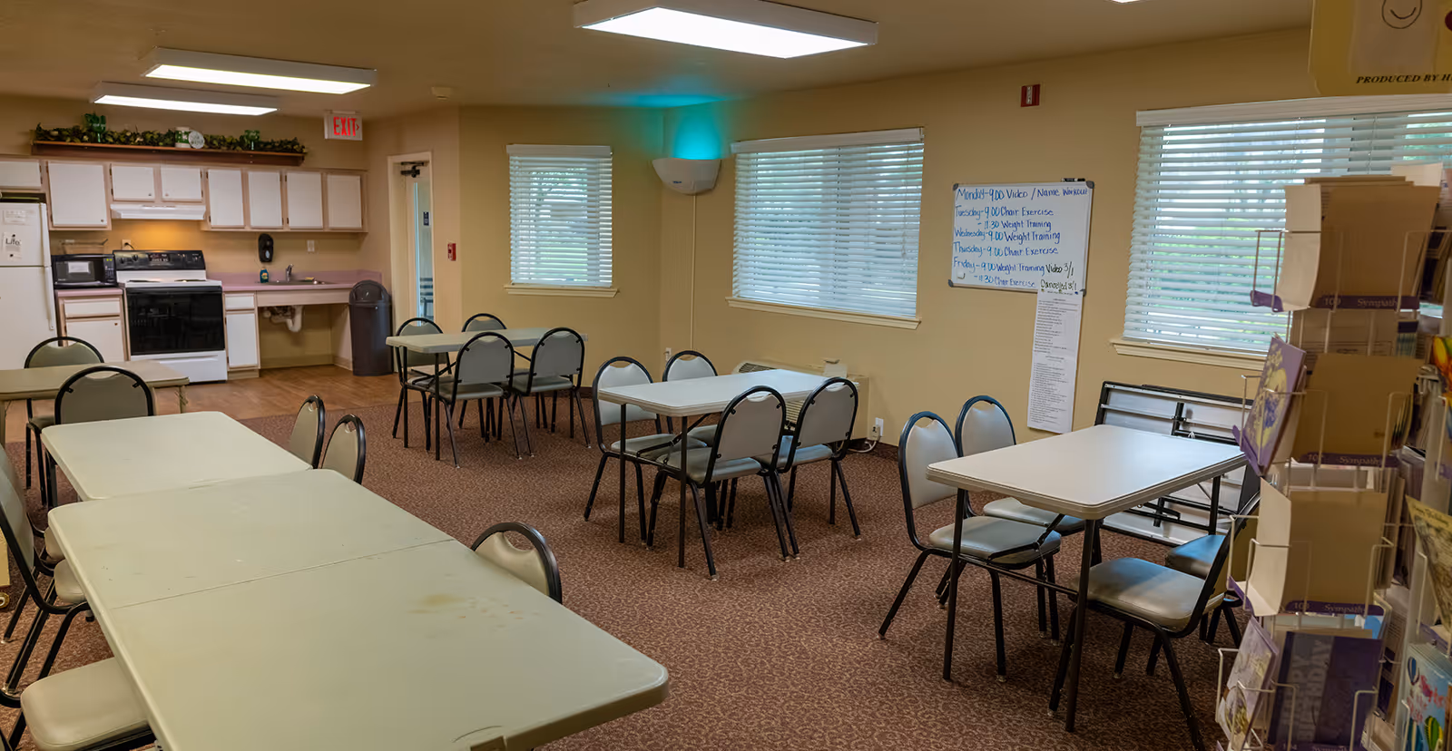 Communal activity/dining room with multiple folding tables and chairs, carpeted floor, and a small kitchenette along the back wall.