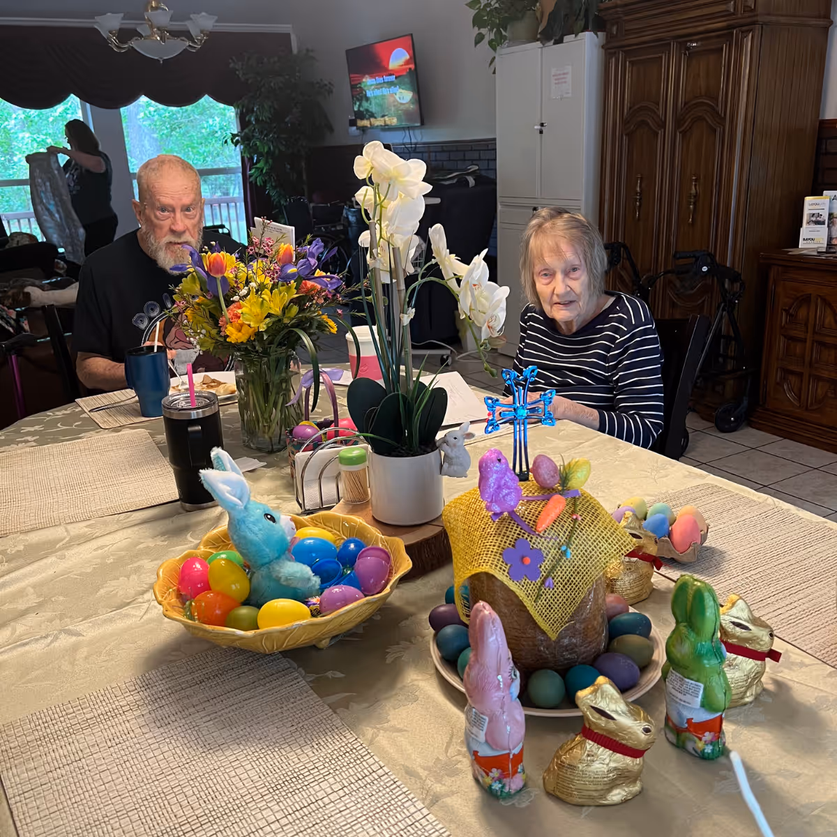 Two elderly residents sit at a dining table decorated with flowers, an orchid, colorful Easter eggs and chocolate bunnies in a senior living dining room.