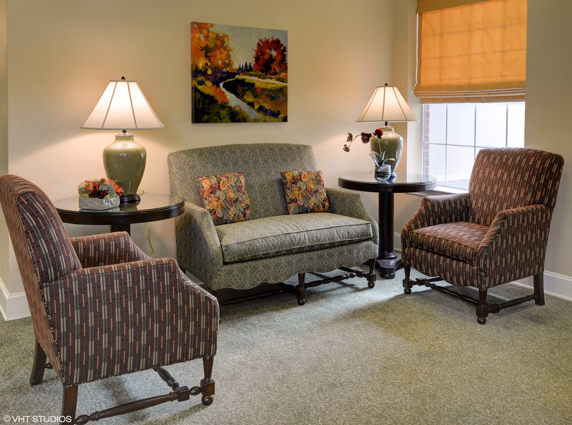 A cozy sitting area with a patterned loveseat adorned with two floral cushions, flanked by two striped armchairs. Each side of the loveseat has a round wooden side table with a green ceramic lamp and decorative items. A colorful landscape painting hangs on the wall above the loveseat, and a window with a beige shade is on the right side.