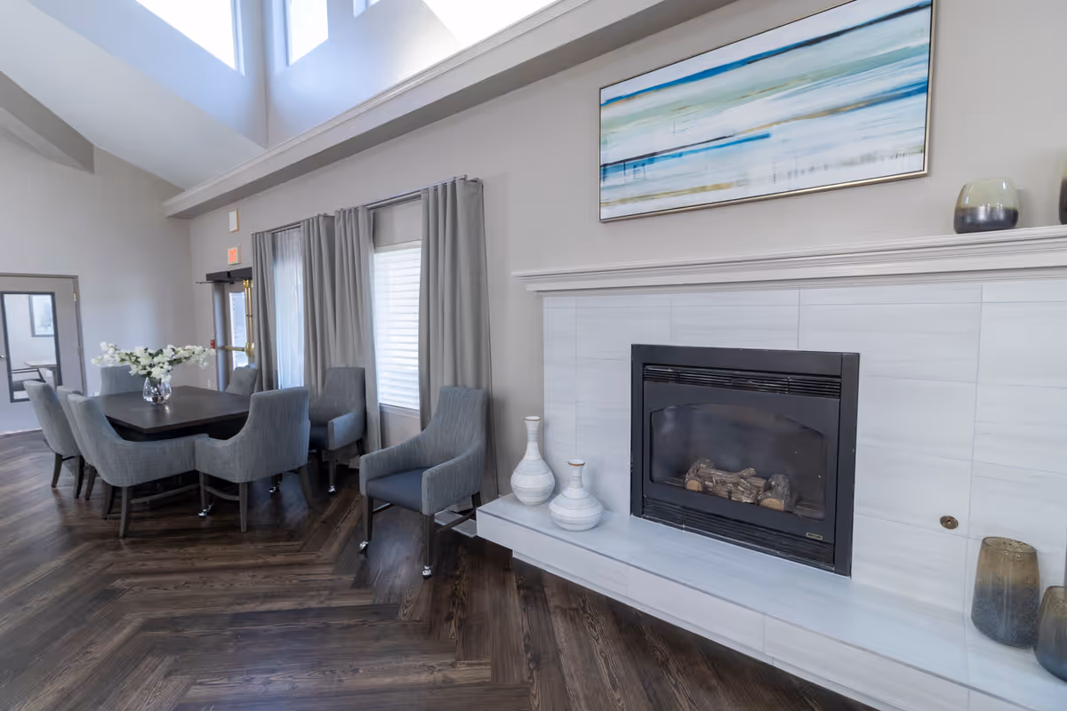 A bright and spacious interior room featuring a modern fireplace with decorative vases on the hearth. To the left, there is a dining area with a dark wooden table surrounded by gray upholstered chairs. Large windows with gray curtains allow natural light to fill the space, and a large abstract painting hangs above the fireplace. The room has dark wood herringbone flooring and high ceilings with skylights.
