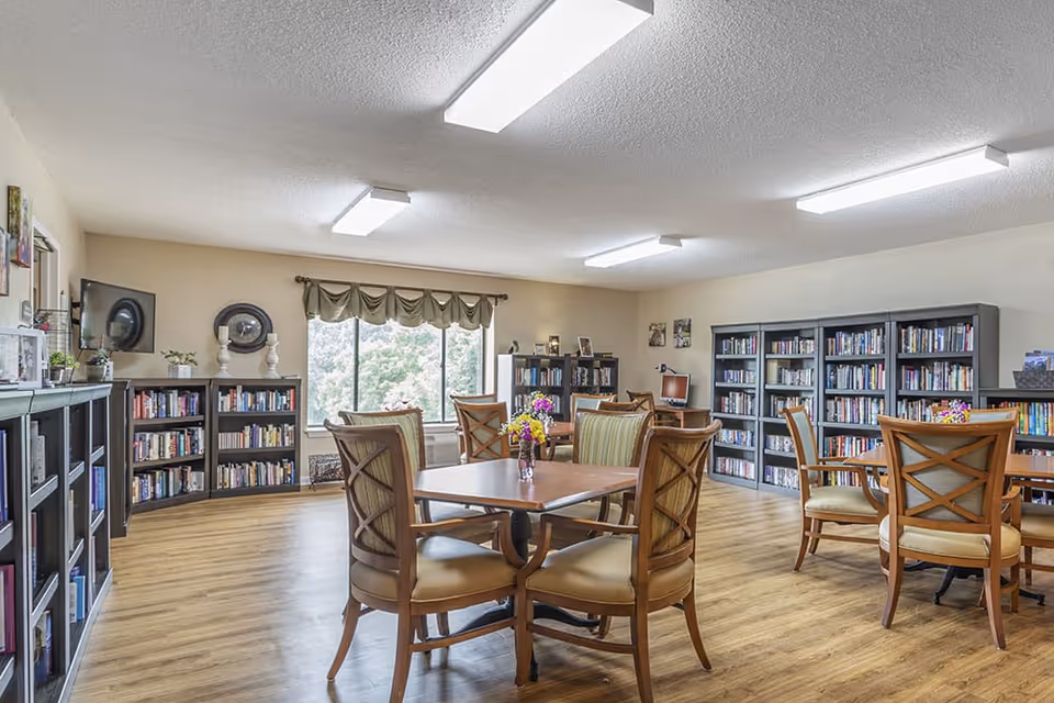 A bright and spacious room with wooden floors, multiple bookshelves filled with books, and several wooden tables with cushioned chairs arranged around them. The room has large windows with a view of greenery outside, and fluorescent ceiling lights providing illumination. Small flower vases are placed on the tables, and a computer is visible on a desk in the background.