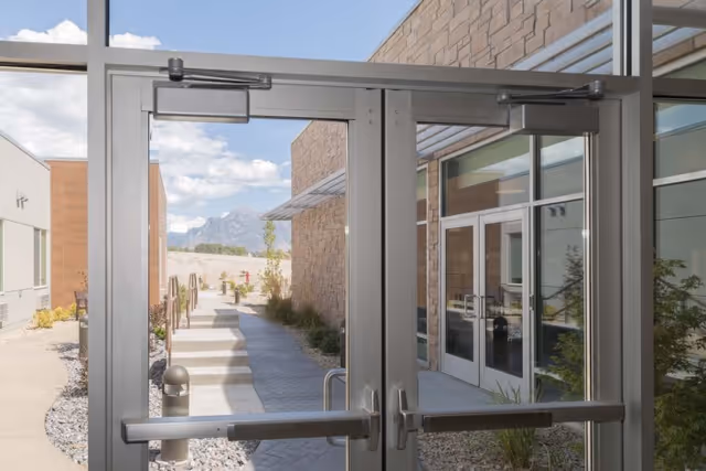 View through glass double doors showing an outdoor walkway with steps, surrounded by modern building walls and landscaping with rocks and plants, under a partly cloudy sky with mountains visible in the distance.