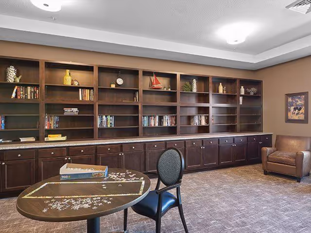 Community lounge with dark wood built-in bookshelves along the back wall, a round table with a jigsaw puzzle and chairs, and a leather armchair.