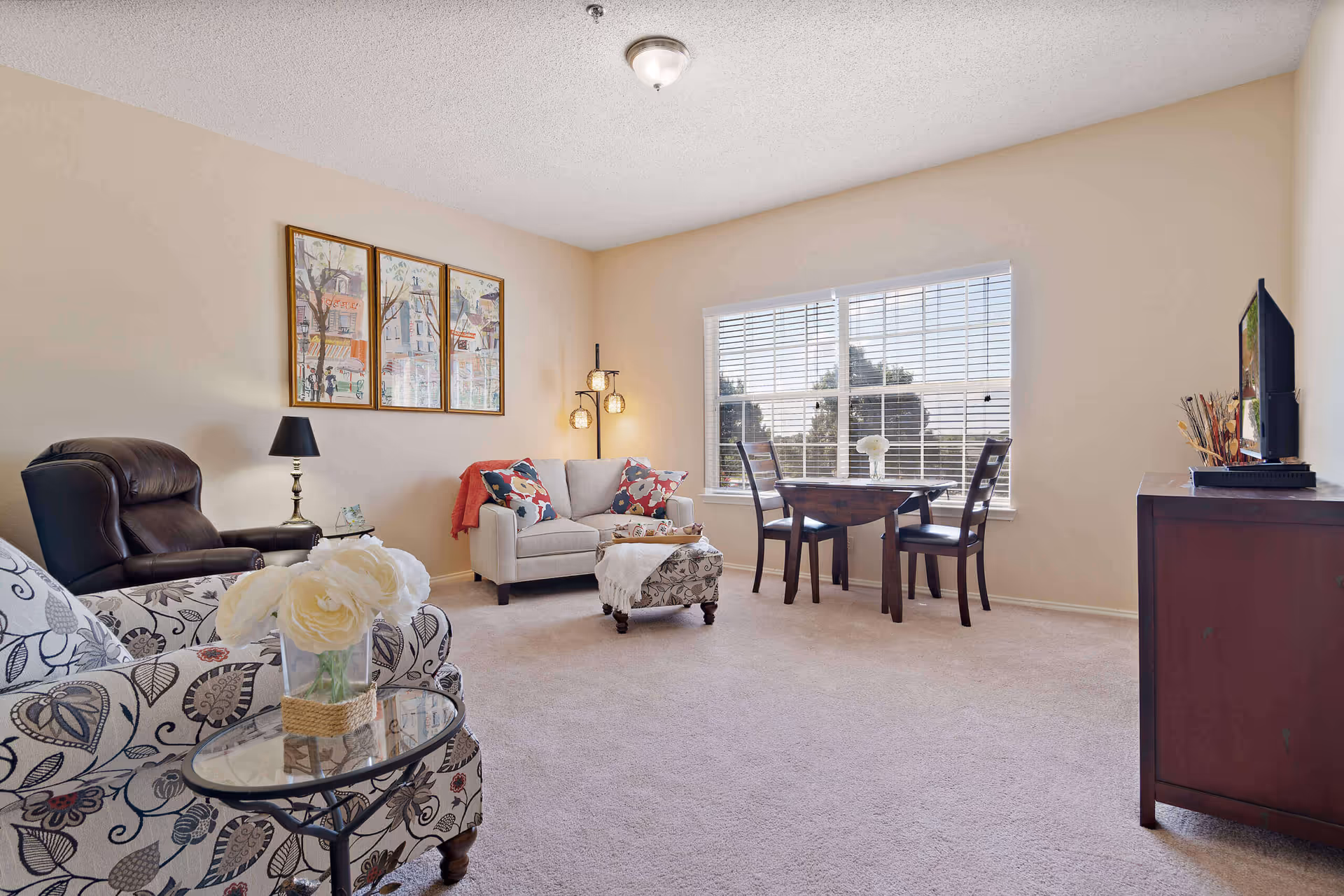 A cozy living room with beige walls and carpet. The room features a patterned armchair with a glass side table holding a vase of white flowers, a dark brown leather recliner, a light gray loveseat with colorful pillows and a throw blanket, and a floral ottoman. There is a wooden dining table with two chairs near a large window with white blinds, allowing natural light to fill the room. A wooden TV stand with a flat-screen TV and decorative items is on the right side. Three framed paintings hang on the wall above the loveseat, and a floor lamp with three globe lights stands in the corner.