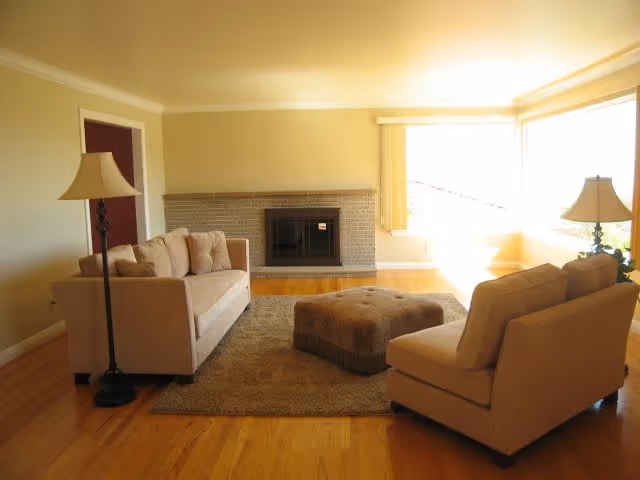 A bright living room with two beige sofas facing each other, a large cushioned ottoman in the center on a brown rug, a floor lamp beside each sofa, and a brick fireplace on the far wall. Large windows on the right side let in natural light.