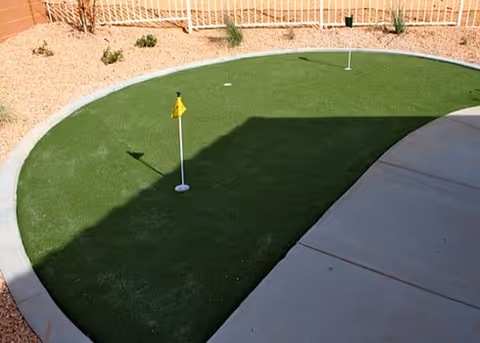 Outdoor putting green with two small flags on a curved artificial turf area surrounded by a concrete walkway and desert landscaping with rocks and sparse plants.