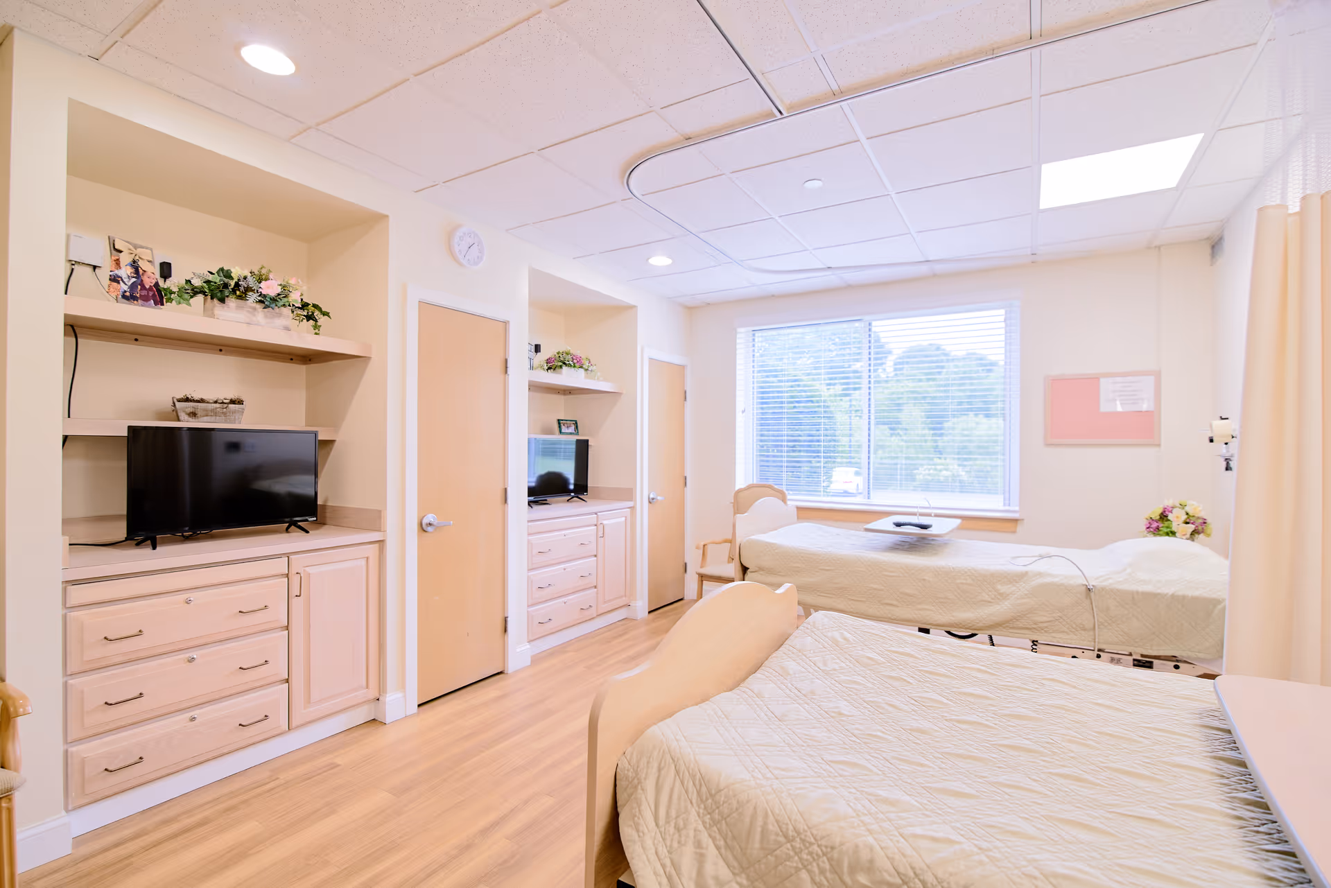 A bright and clean senior living facility bedroom with two beds covered in beige quilts, a wooden chair, and a large window with blinds letting in natural light. The room has light wood flooring, built-in shelves with a TV, decorative plants, and a clock on the wall.