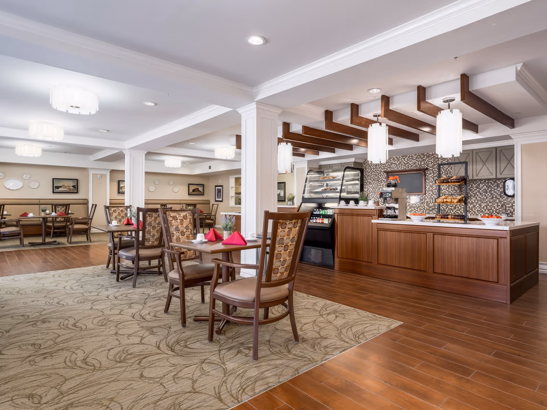 A bright and spacious dining area in a senior living facility with wooden tables and chairs, some tables set with red napkins and white cups. The room features a serving counter with pastries and a coffee machine, decorative ceiling beams, and modern pendant lights. The floor is a combination of wood and patterned carpet, and the walls are adorned with framed pictures and decorative plates.
