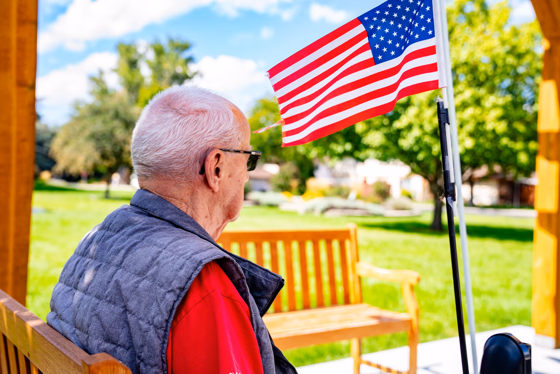 An elderly man with white hair and glasses sits on a wooden bench outdoors, wearing a red shirt and a gray vest. In front of him is an American flag mounted on a pole. The background shows green grass, trees, and a bright blue sky with some clouds.