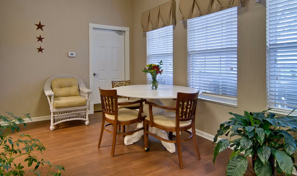 A cozy interior room with a round white table surrounded by four wooden chairs with cushioned seats. A vase with flowers is placed on the table. There is a white wicker chair with a yellow cushion against the wall, which has three decorative star ornaments. The room has large windows with blinds and beige valances, and there are green plants on the floor. The floor is wooden.