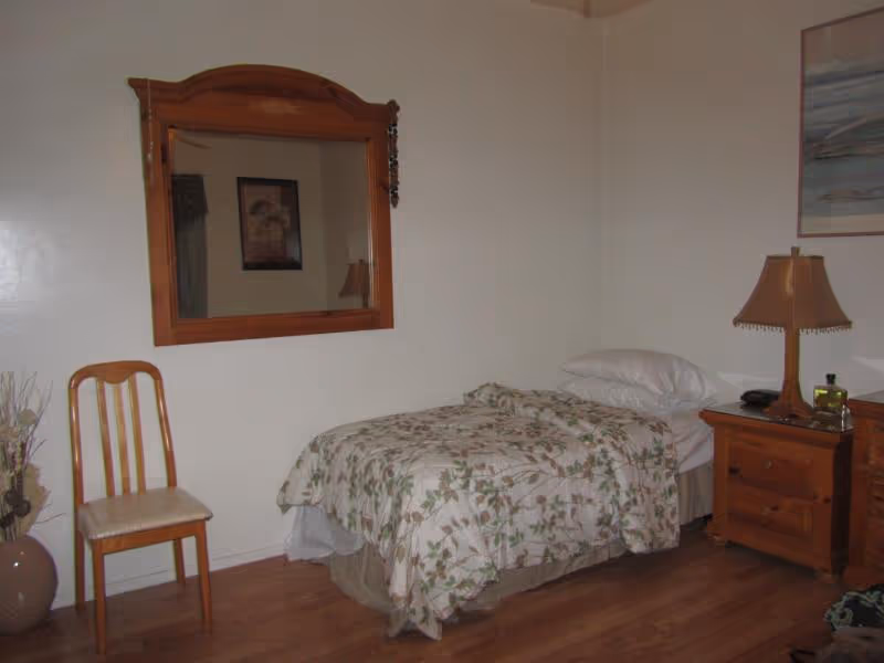 A small bedroom with a single bed covered in a floral patterned bedspread. Next to the bed is a wooden nightstand with a lamp and some items on it. A wooden chair is placed against the wall under a large wooden-framed mirror. The floor is wooden, and the walls are plain white with a framed picture hanging above the nightstand.