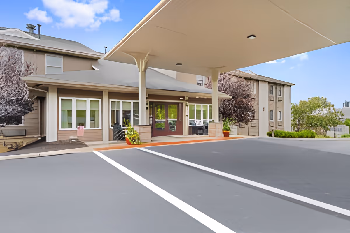 Entrance area of a senior living facility with a covered drop-off zone, large windows, and a paved driveway with white lines. The building is surrounded by some trees and greenery under a blue sky with clouds.