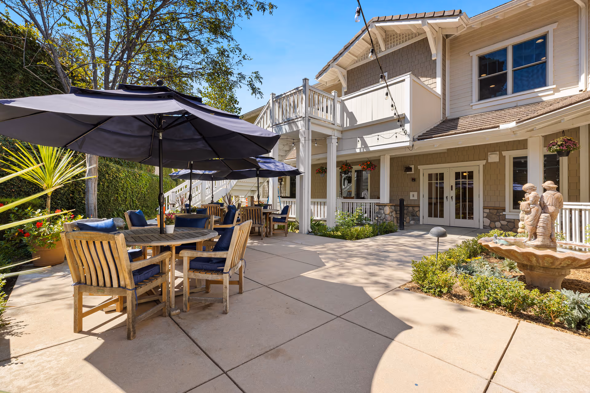 Outdoor patio area at Ivy Park at Wood Ranch with wooden tables and chairs under large navy blue umbrellas, surrounded by plants and greenery. The building exterior features beige siding, white trim, hanging flower pots, and a stone fountain with a sculpture of three children.