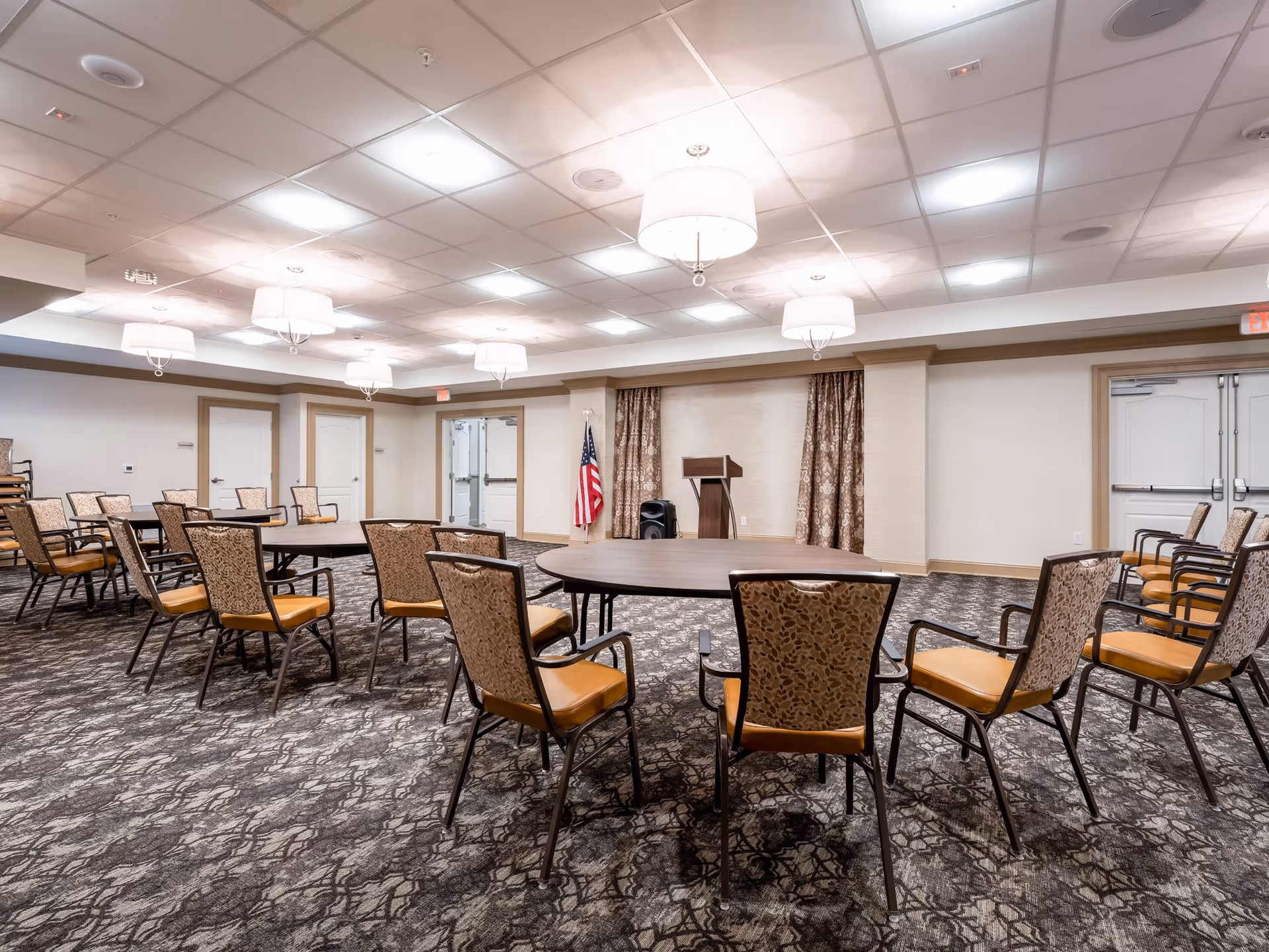 A spacious meeting or event room with round tables and patterned chairs arranged around them. The room has a carpeted floor with a dark, intricate pattern, white walls with beige trim, and a ceiling with recessed lighting and hanging light fixtures. At the far end, there is a podium with an American flag beside it and double doors on either side.
