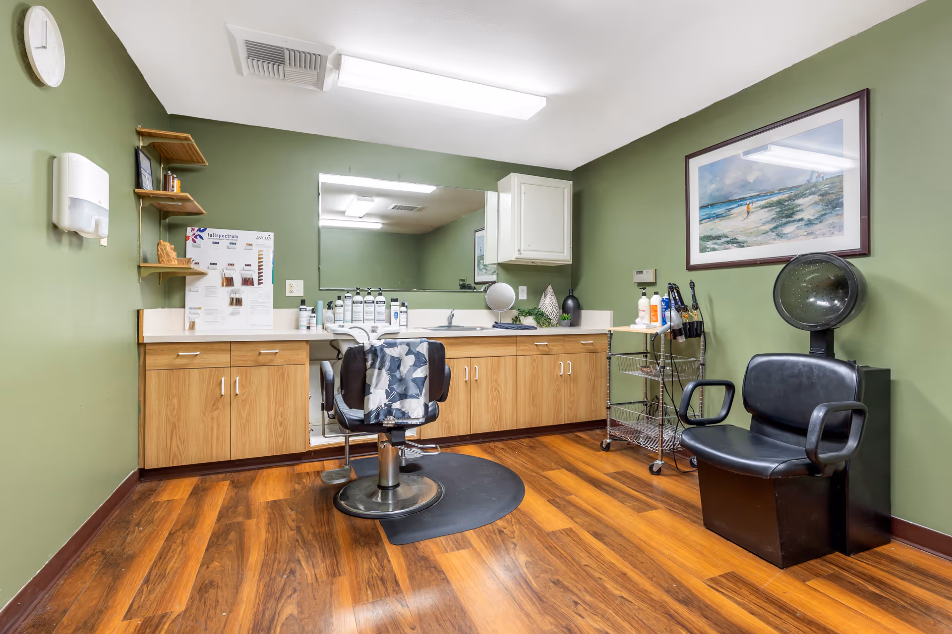 Interior of a hair salon room with green walls and wooden flooring. The room features a large mirror above a counter with various hair care products, a black salon chair with a floral cape draped over it, a black hair dryer chair, a small rolling cart with more hair products, and a framed beach landscape painting on the wall.