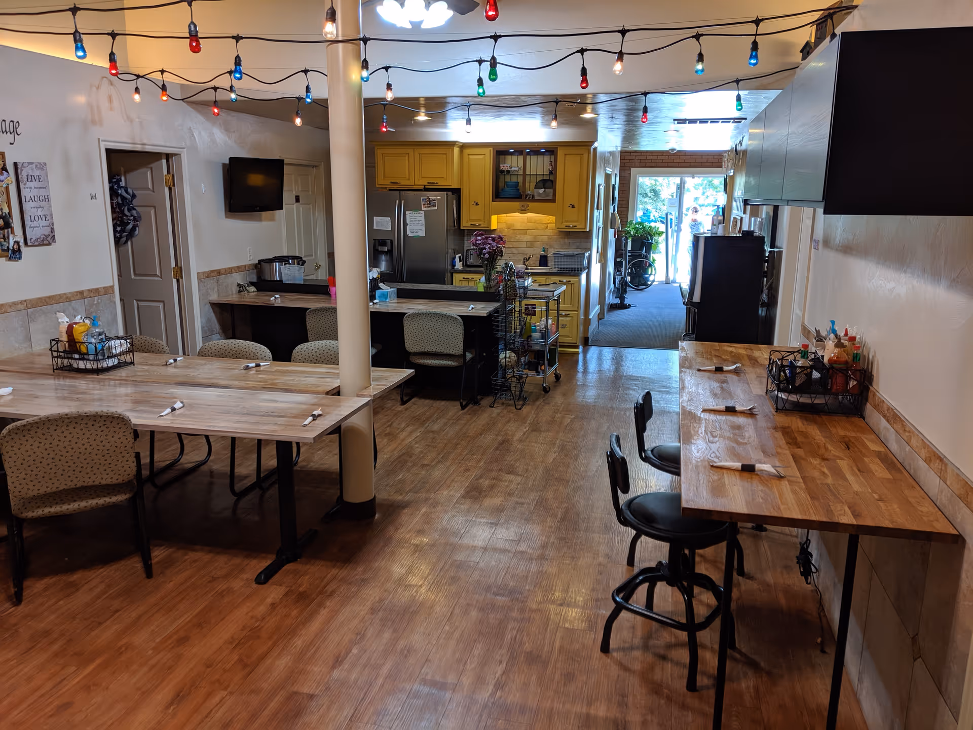 Communal dining area with wooden tables and chairs, a counter with stools, and a kitchen in the background decorated with string lights.