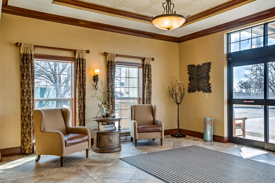 Sunlit lobby with two upholstered armchairs flanking a round wooden side table, decorative wall art, windows and a glass entrance door.
