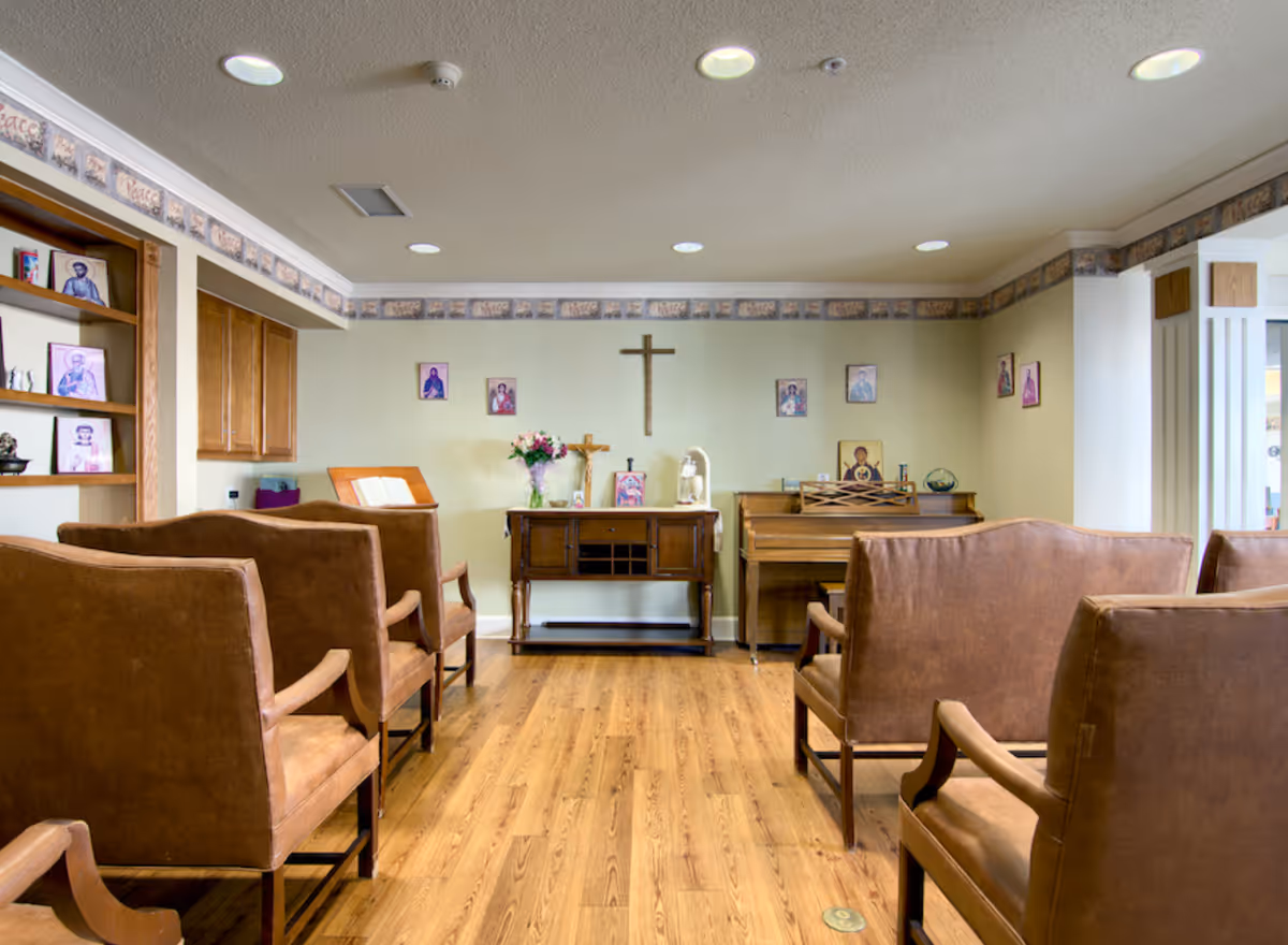A small chapel or prayer room with wooden chairs arranged in rows facing a wall with religious icons, a wooden cross, a piano, and a cabinet with flowers and religious items. The room has wooden flooring and soft lighting from ceiling lights.