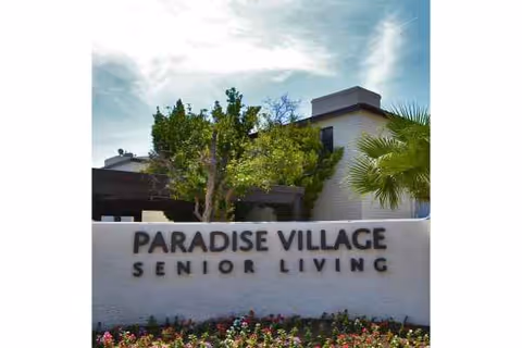 Exterior view of Paradise Village Senior Living with a white wall, sign, trees, and building.