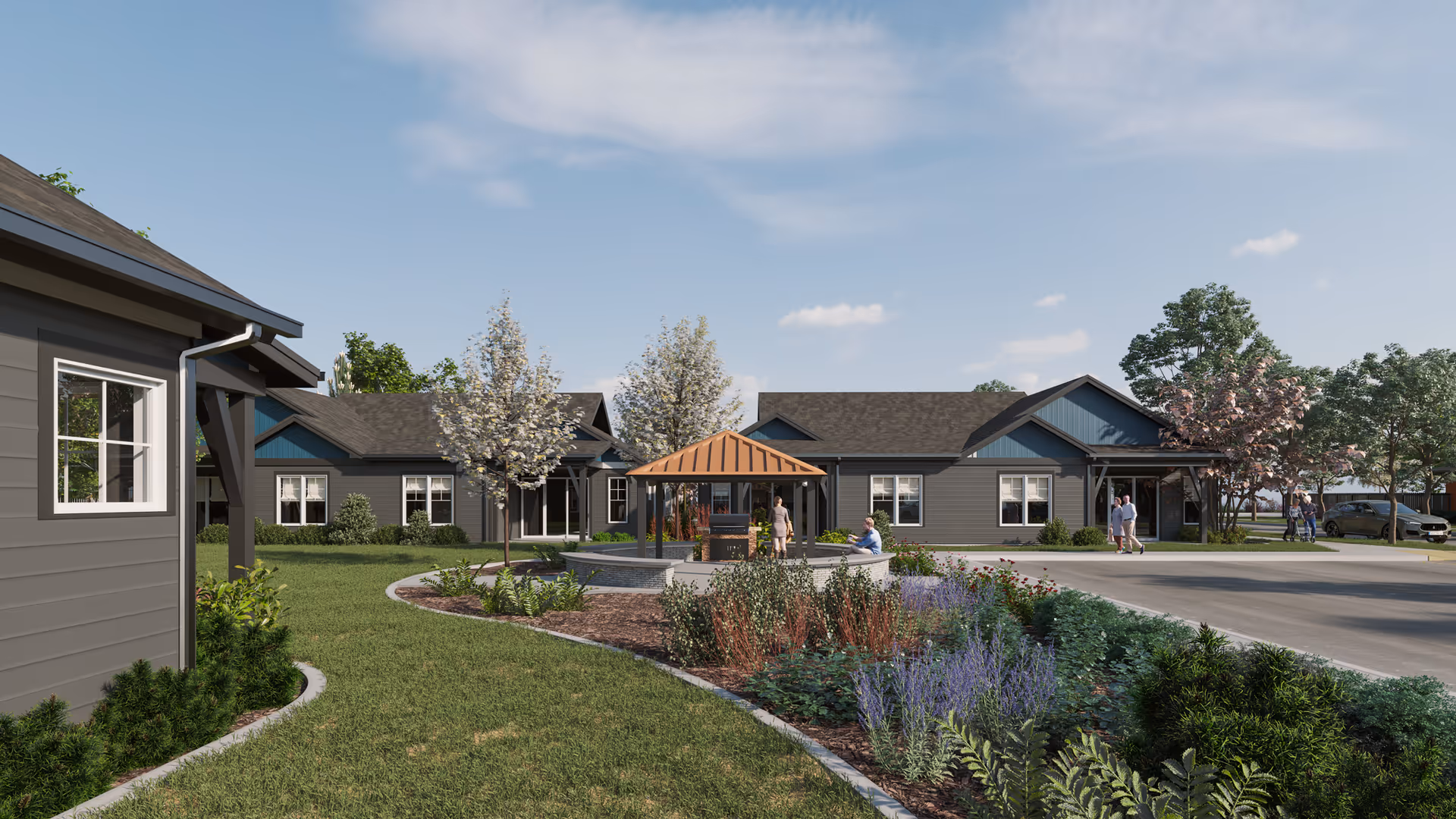 Outdoor view of a senior living facility with single-story buildings, landscaped garden beds with various plants and flowers, a circular seating area with a wooden pergola, and several people walking and sitting outside under a partly cloudy sky.