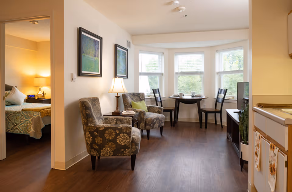 A cozy senior living apartment interior showing a small sitting area with two patterned armchairs and a side table with a lamp. In the background, there is a round dining table with two chairs near three large windows letting in natural light. To the left, a bedroom with a bed and nightstand with a lamp is partially visible. To the right, part of a kitchen counter and cabinets can be seen.