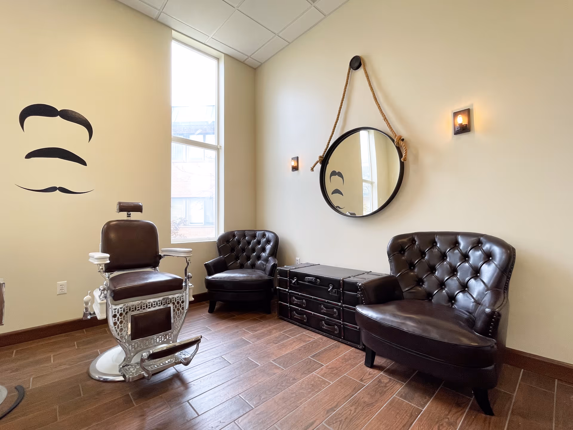 A stylish room with a vintage barber chair, two dark brown tufted leather chairs, a round mirror hanging on the wall with a thick rope, and a wall decoration of various mustache styles. The room has a large window letting in natural light and wooden flooring.