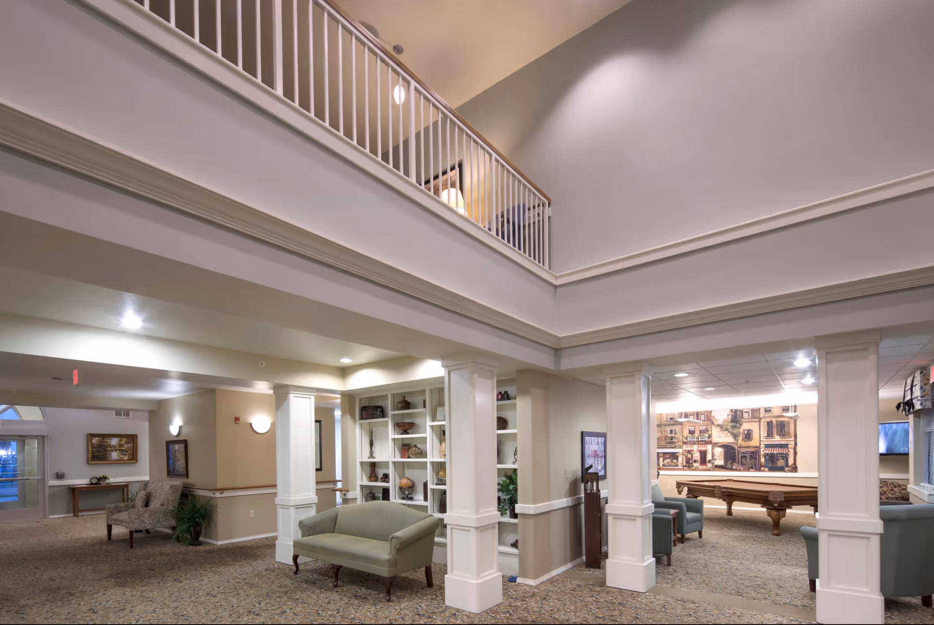 Spacious interior common area of an assisted living facility with high ceilings and a second-floor balcony. The area features a green upholstered bench, built-in white shelves with decorative items, and a seating area with armchairs around a pool table. The walls are painted in neutral tones and decorated with framed artwork.