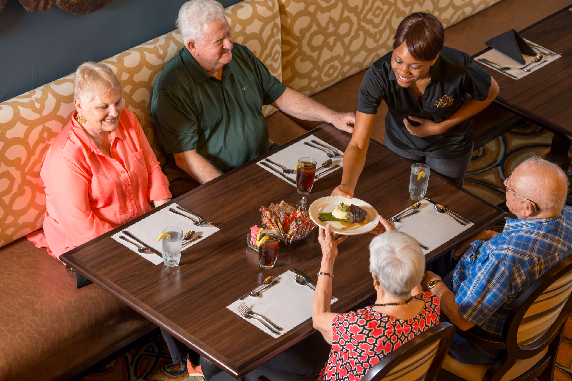 A waitress serving a plate of food to an elderly woman seated at a dining table with three other elderly people in a senior living facility dining room.