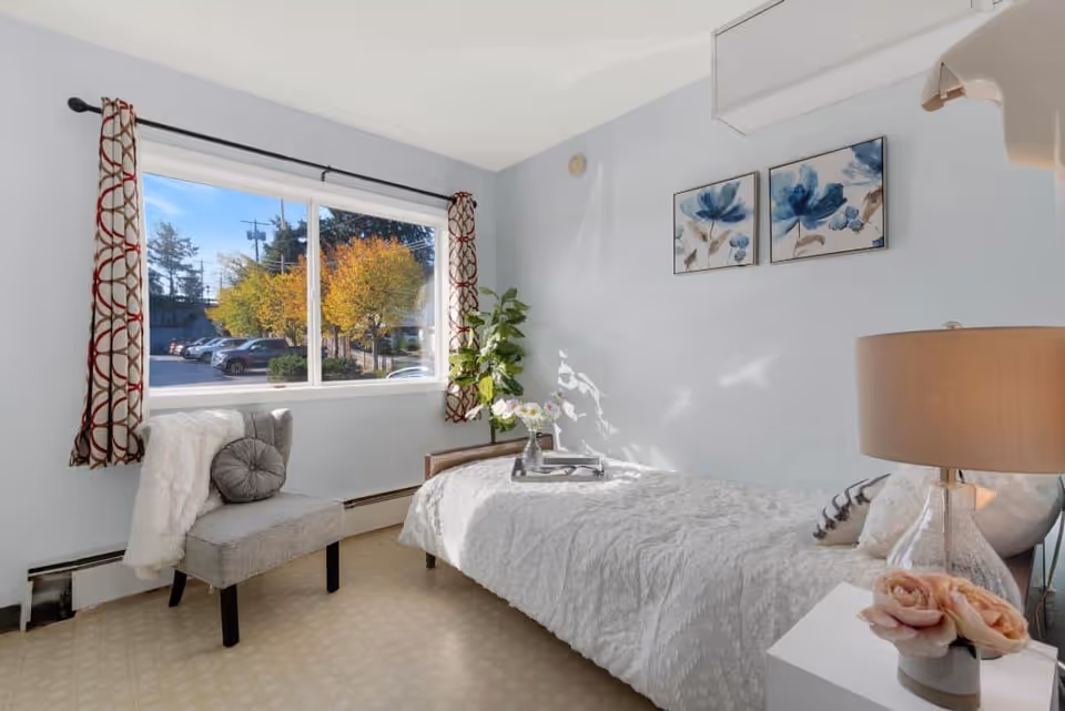 A bright and cozy bedroom with a single bed covered in a white textured bedspread. Next to the bed is a white nightstand with a clear glass lamp and a small vase with pink flowers. A gray upholstered chair with a round pillow and a white throw blanket is placed near a large window with patterned curtains. Outside the window, trees with autumn-colored leaves and parked cars are visible. Two blue floral paintings hang on the light blue wall above the bed.