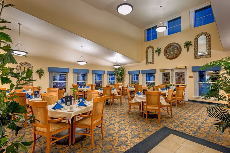 Spacious, well-lit dining room with multiple set tables and chairs, blue napkins, plants, and a large clock on the wall beneath high ceilings.