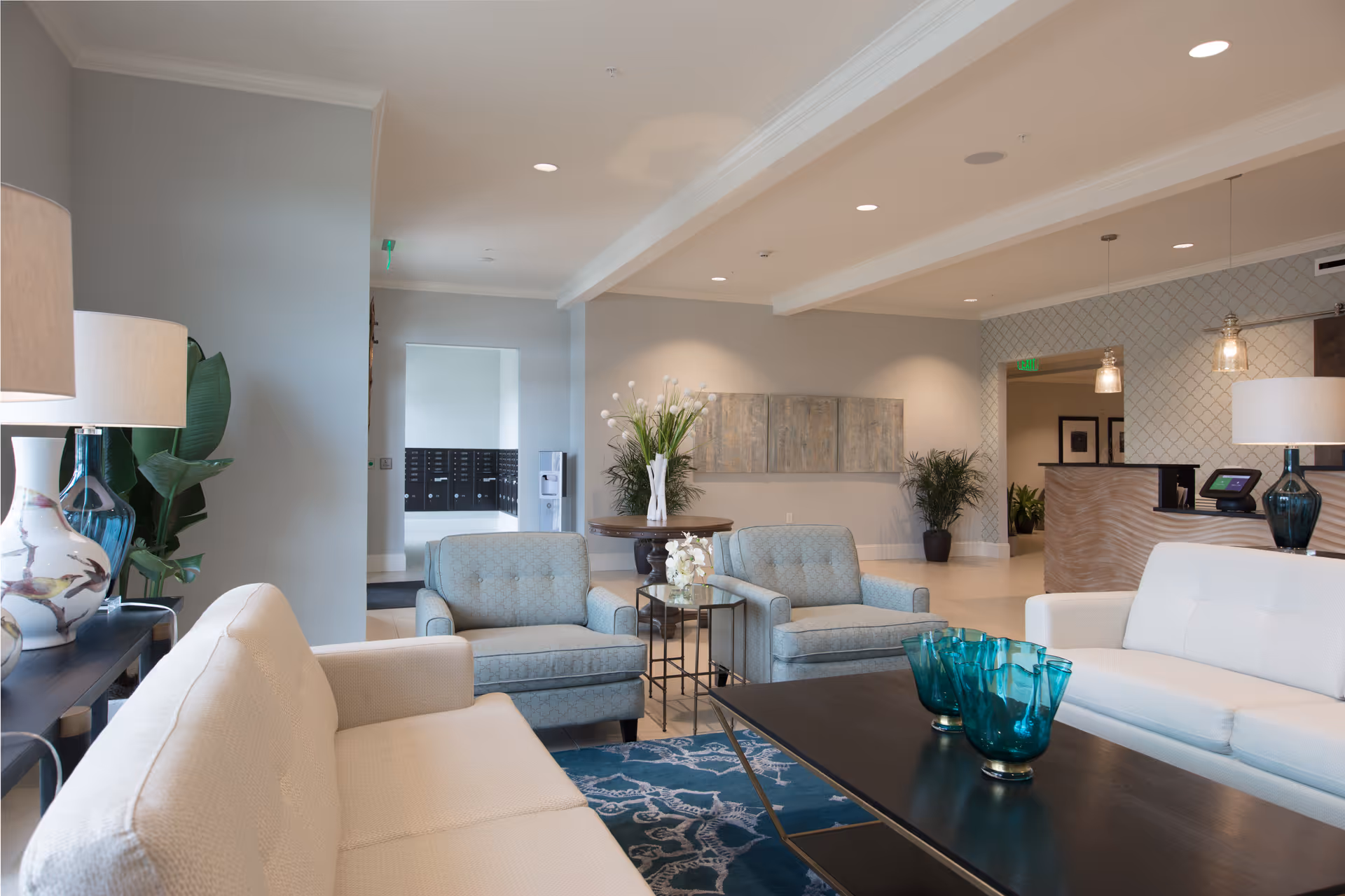 A bright and spacious living room area in a senior living facility featuring two light blue armchairs, two white sofas, a dark coffee table with blue glass vases, a patterned blue rug, decorative lamps, plants, and a reception desk in the background.