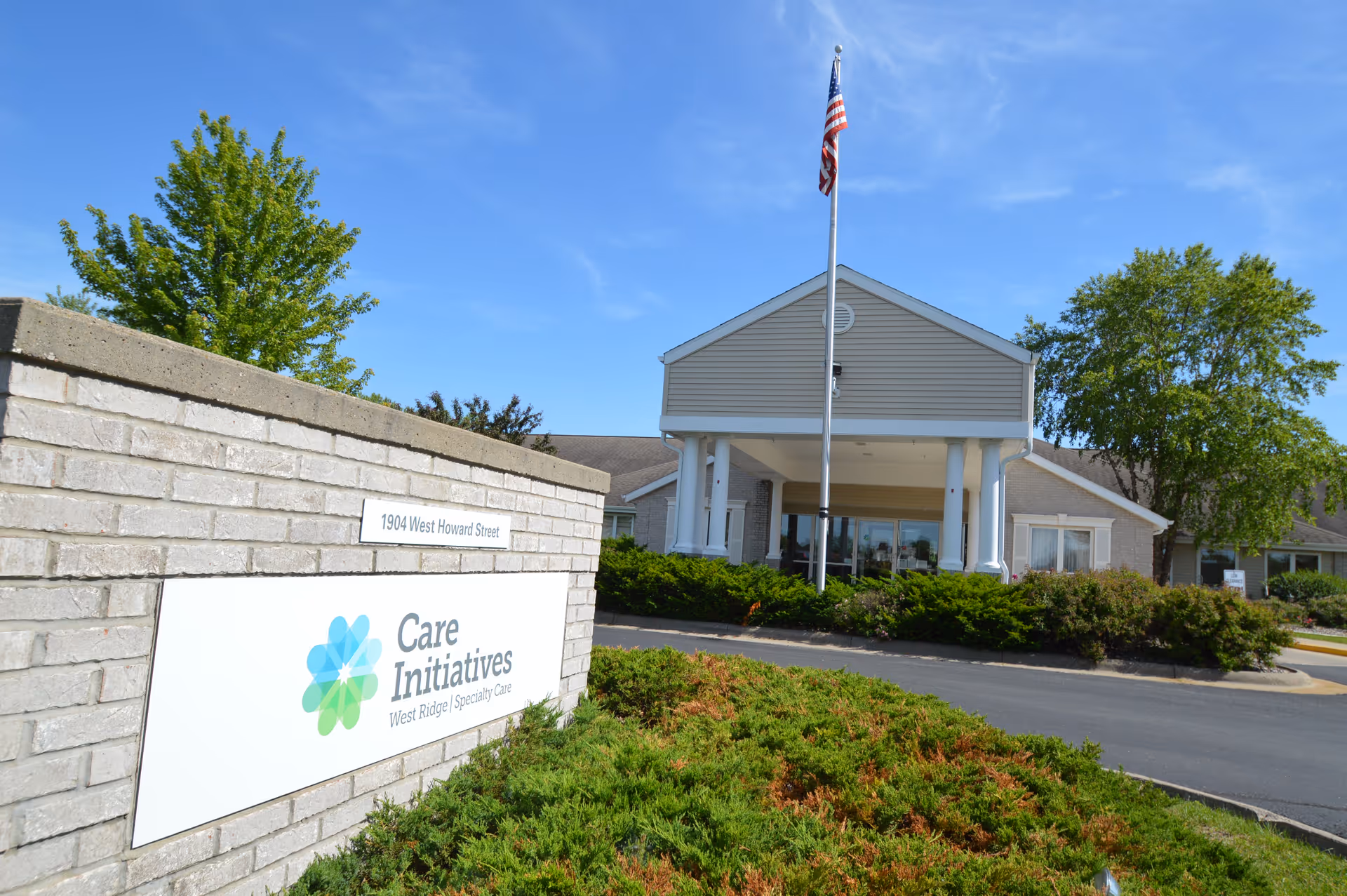 Exterior view of West Ridge Specialty Care facility with a brick sign in the foreground displaying 'Care Initiatives West Ridge Specialty Care' and the address '1904 West Howard Street'. The building has a covered entrance with white columns and an American flag flying on a flagpole in front. Green bushes and trees surround the area under a clear blue sky.