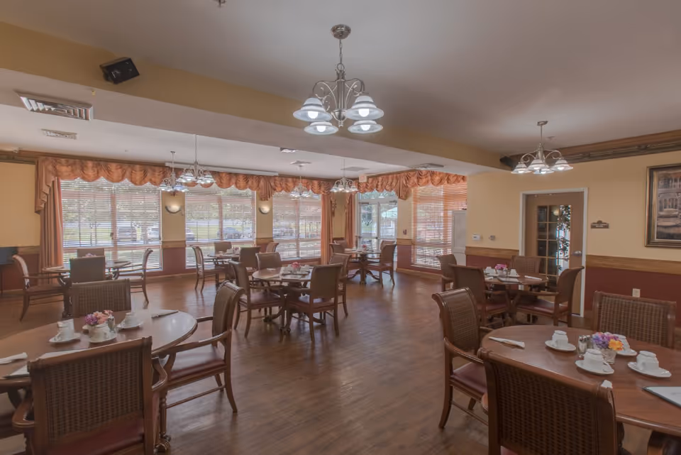 A dining room with multiple round wooden tables and chairs arranged neatly. Each table is set with white cups, saucers, napkins, and small flower arrangements. Large windows with blinds and decorative valances allow natural light to fill the room. The floor is wooden, and there are several ceiling light fixtures providing additional lighting. A door and a framed picture are visible on the right wall.