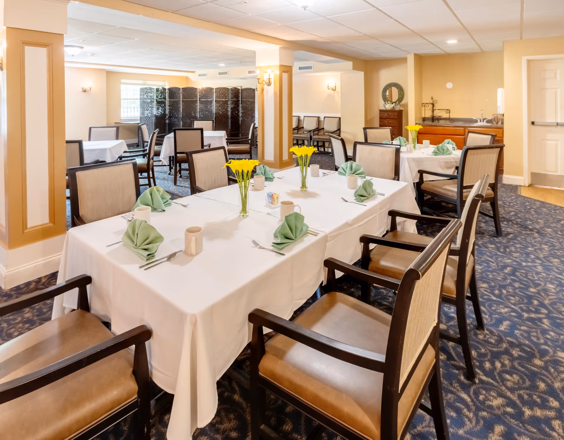 Bright dining room with tables set with white tablecloths, folded green napkins, yellow flower centerpieces, and wooden chairs.