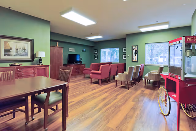 A spacious common area with green walls and wood flooring featuring a dining table with chairs, a seating area with armchairs and a TV, and a red popcorn machine on wheels near the window.