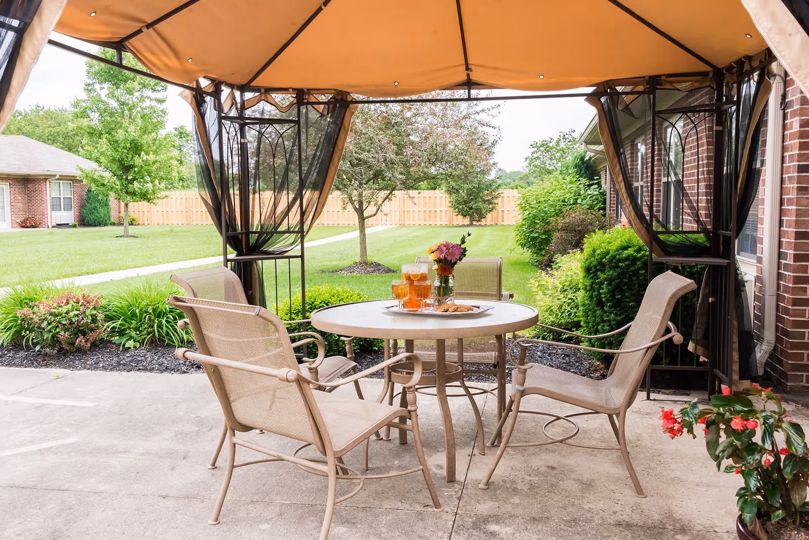 Outdoor patio with a round table set with drinks and flowers under a canopy, surrounded by chairs and a lawn with a fence in the background.