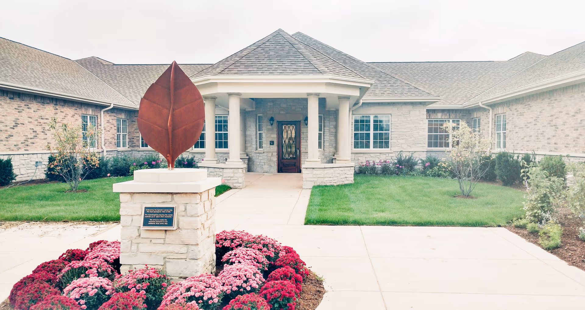 Front exterior view of Franklin Place Memory Care facility featuring a stone building with a covered entrance supported by columns, a pathway leading to the door, a landscaped garden with green grass, bushes, and a flower bed with pink and red flowers surrounding a stone pedestal topped with a large metal leaf sculpture.