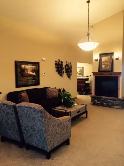 A cozy living room with a dark sofa and two patterned armchairs around a coffee table with a plant. The room features a fireplace with a framed picture above it, wall-mounted decorative pieces, and warm lighting from a hanging ceiling lamp and wall sconces.