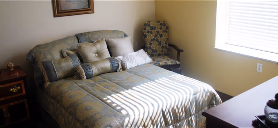 A cozy bedroom with a neatly made bed featuring multiple decorative pillows and a patterned bedspread. There is a wooden nightstand with a small decorative item on it to the left of the bed, a cushioned armchair with a floral pattern in the corner, and a window with blinds letting in sunlight on the right side.