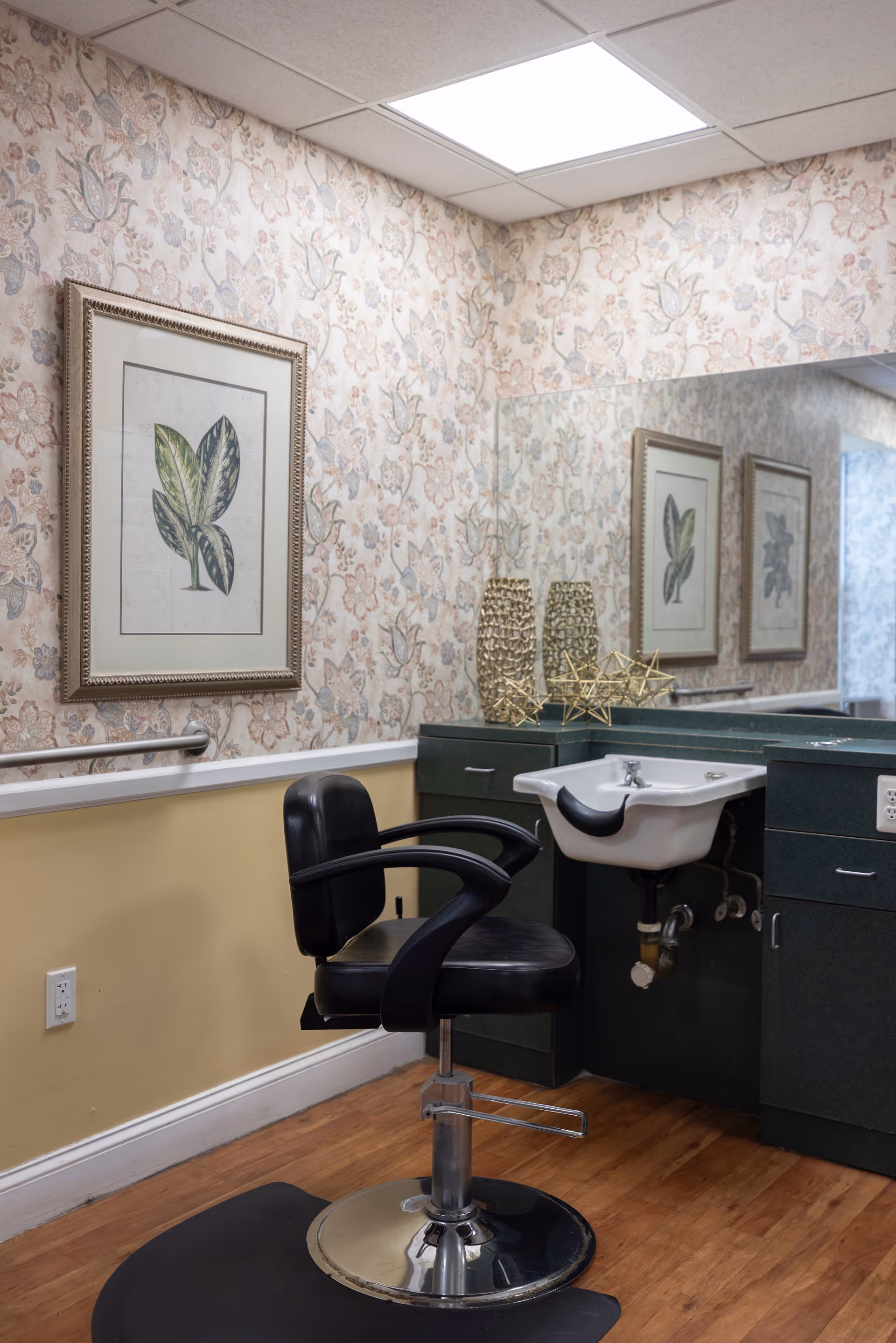 Interior view of a salon area in an assisted living facility featuring a black salon chair in front of a white sink with a black neck rest. The walls have floral patterned wallpaper with a framed botanical print. There is a large mirror above the sink reflecting the room, and decorative gold geometric sculptures are placed on the countertop.