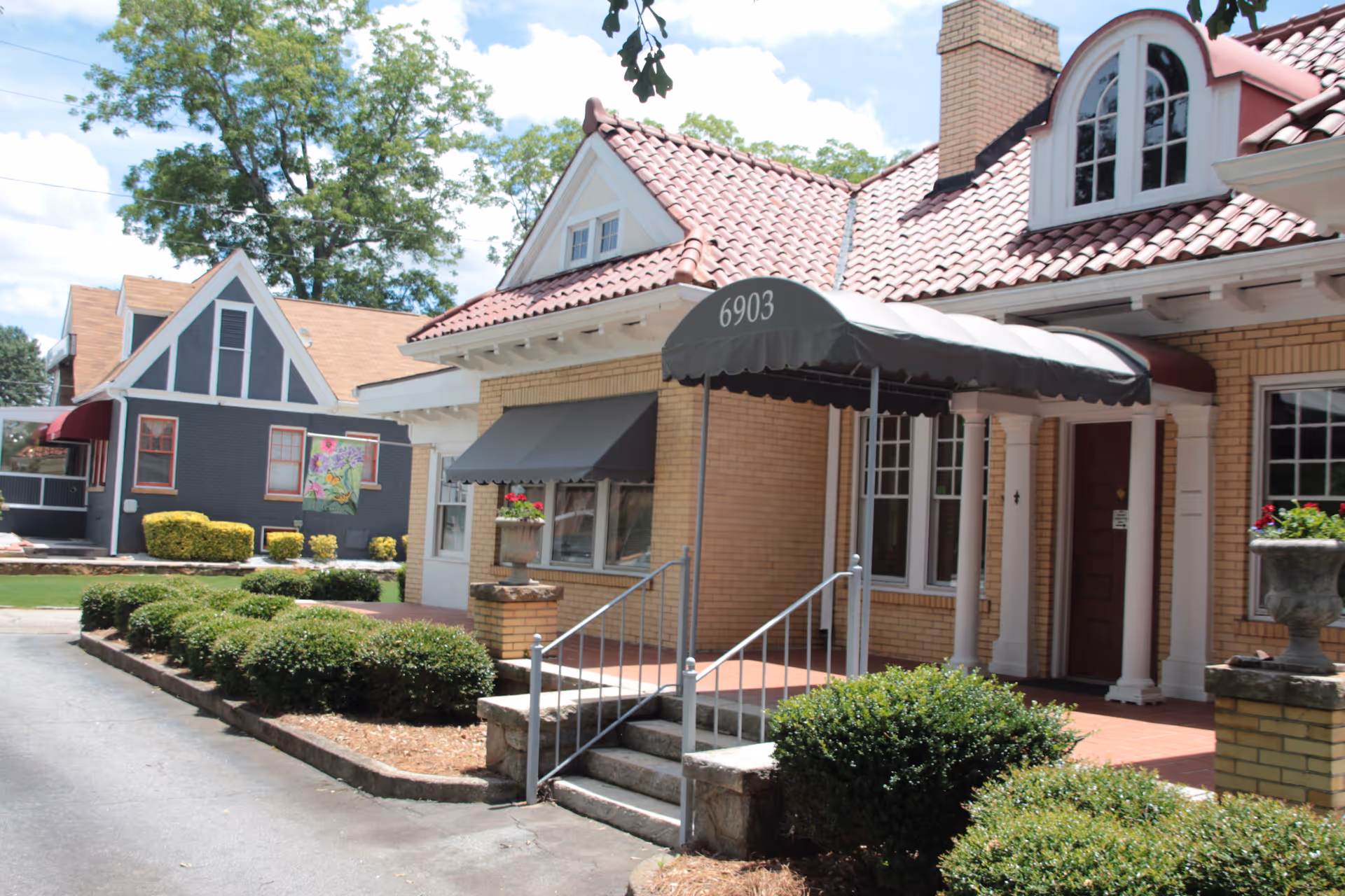 Exterior view of a brick building with a red tile roof and a black awning displaying the number 6903. The entrance has steps with metal railings and is flanked by bushes and planters with flowers. Another house with a dark exterior and white trim is visible in the background under a partly cloudy sky.