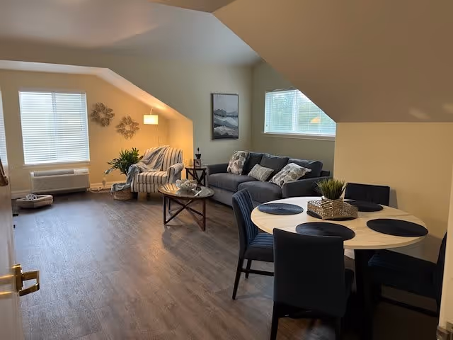 Open living and dining area with a sofa, armchair, coffee table, and round dining table under a sloped ceiling.