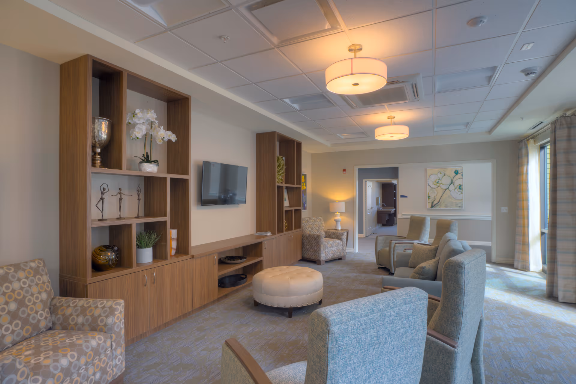 Bright communal living room with upholstered armchairs, an ottoman, built-in shelving and a wall-mounted TV.